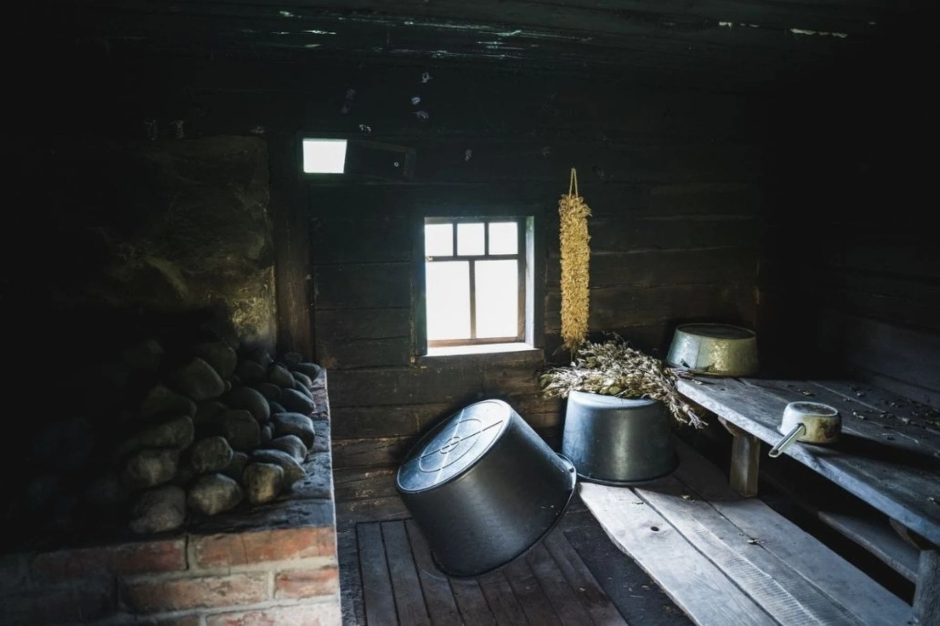 Dark interior of a traditional Estonian smoke sauna with heated stones, dried herbs, and metal washbasins