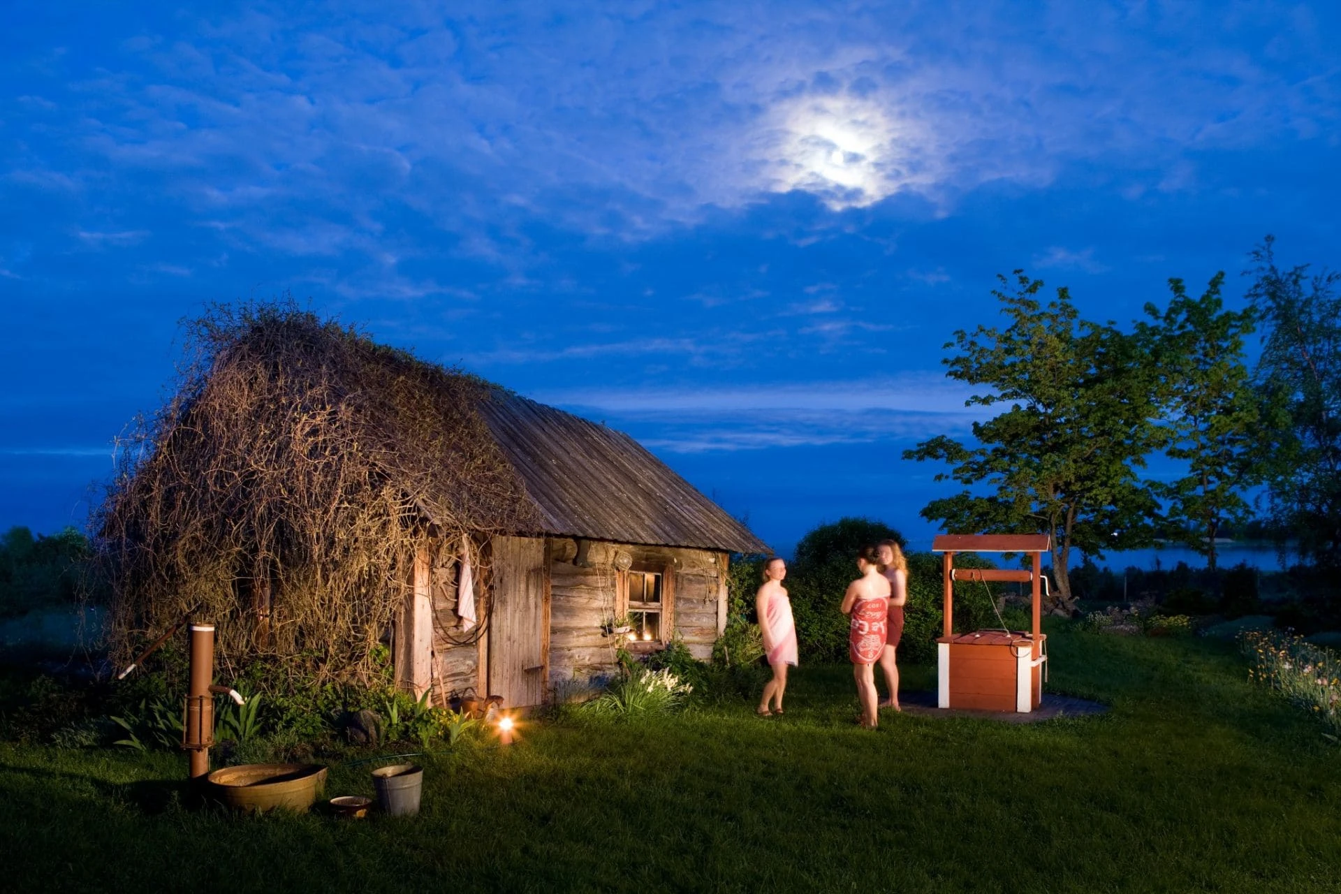Three women in towels standing by a well next to a traditional Estonian sauna under a full moon