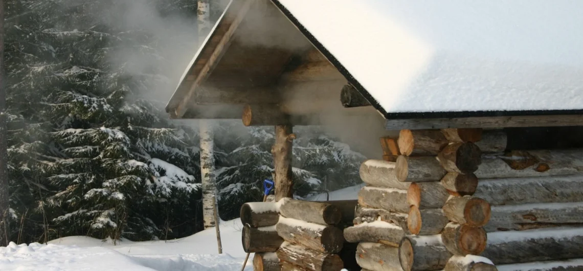 Traditional Estonian log smoke sauna covered in snow with steam rising from the roof in a winter forest